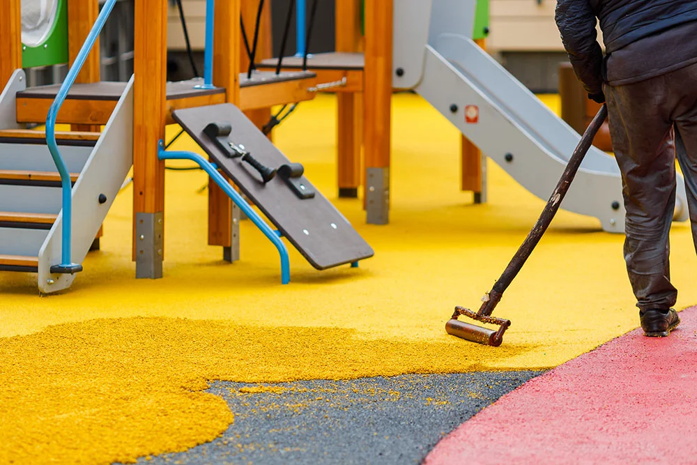 Worker laying down rubber mulch surfacing with a roller on a children's playground.